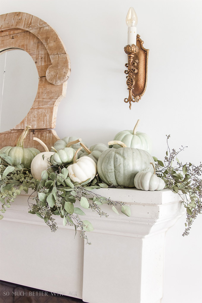 A white mantelpiece with on top 10 pale green and white pumpkins of different sizes, sitting on top of a bed of greenery.